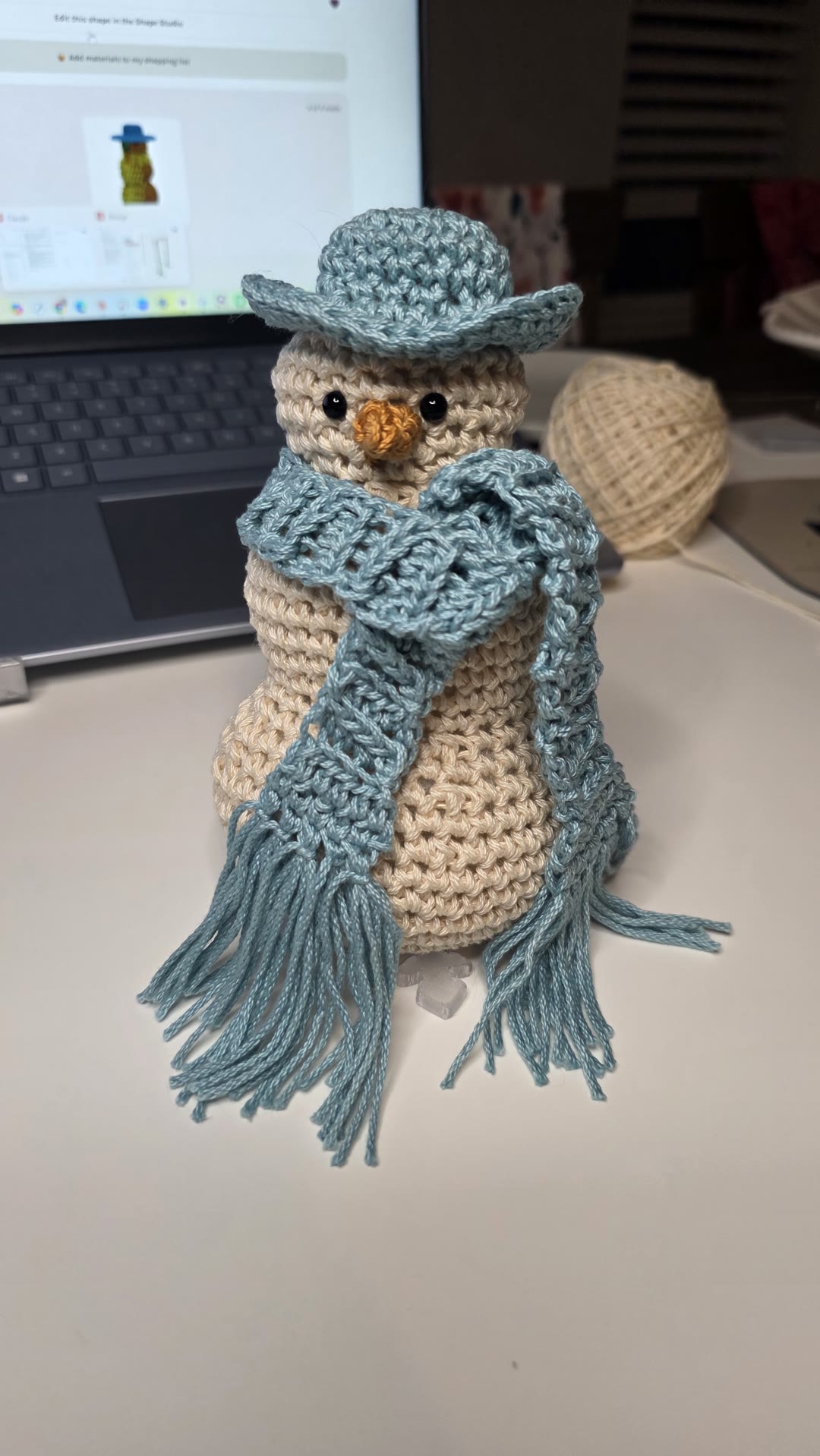 A crocheted cream snowman with black safety eyes, an orange nose, a blue-green brimmed hat and a fringed scarf, standing on a white desk in front of a laptop showing his 3D design in Crochet Bobble